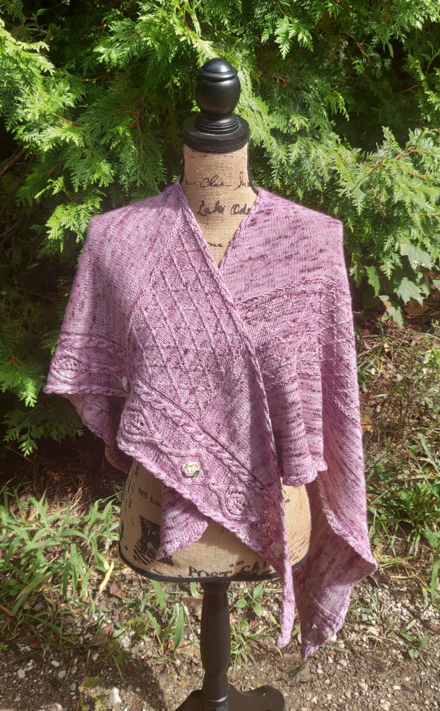 A mottled mauve shawl with purple speckles is draped over a burlap covered mannequin. One edge has a border of two twist cables and a central motif of cable framed diamonds with alternating lace leaves and crocheted roses with a small leaf. There is a criss-cross trellis section visible on each side of the shawl.